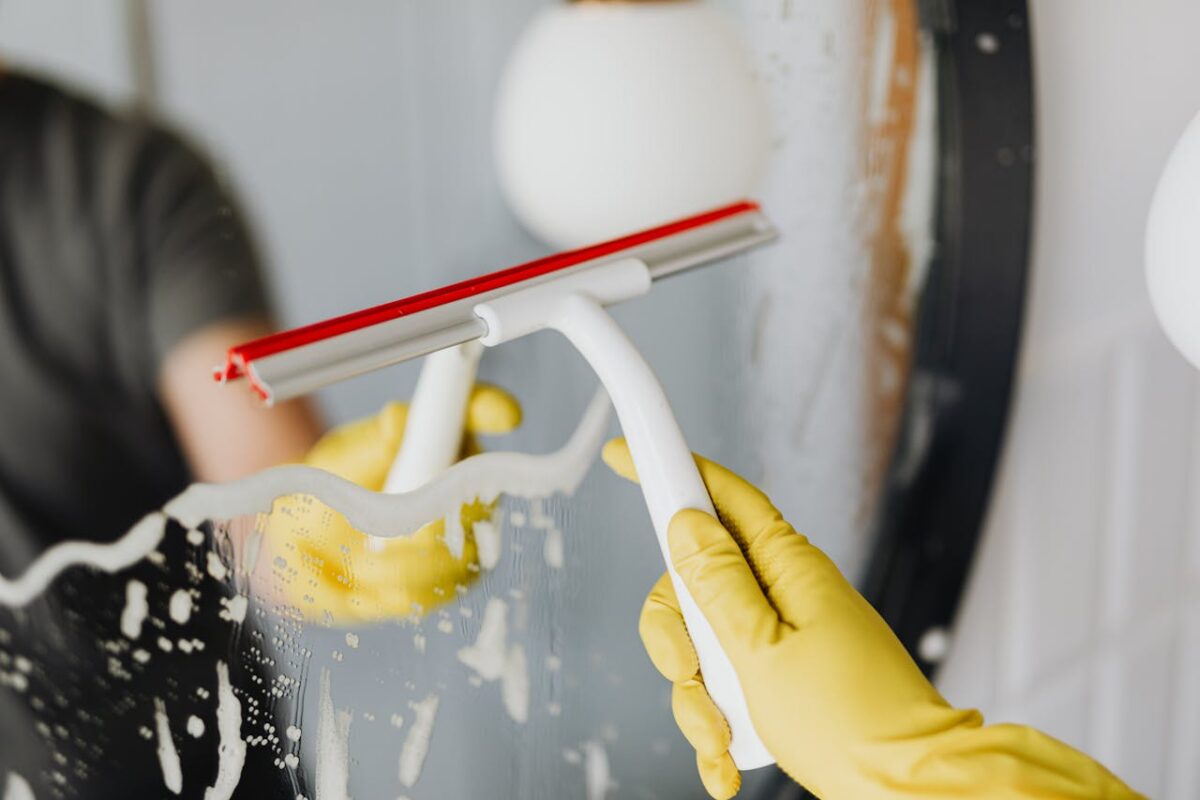 Faceless person in yellow rubber gloves using scraper to remove soapy water from mirror in bathroom